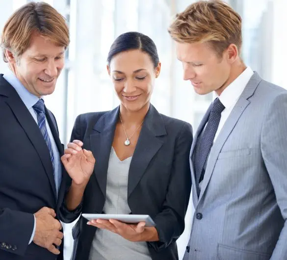 Three business professionals, two men and one woman, gather around a tablet, appearing to discuss content on the screen. The woman in the center holds the tablet and points to it, while the men on either side look on. All are dressed in business attire.